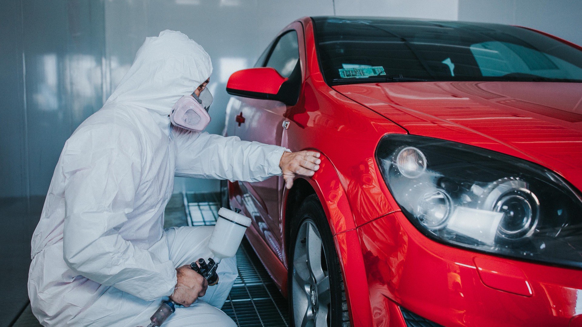 Pintor con traje y mascarilla trabajando el costado de un automóvil rojo dentro de la cabina de pintura