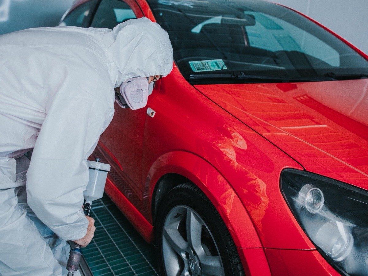 Pintor con traje y mascarilla trabajando el capó de un automóvil rojo en cabina
